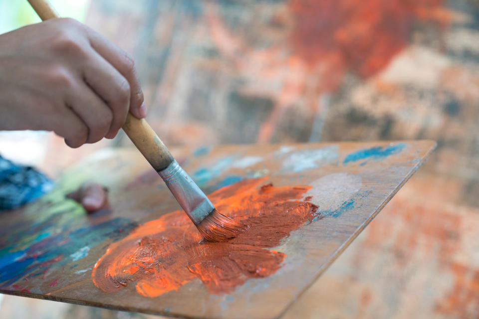 Closeup shot of a painter's hands, picking up paint on a brush from a well-loved palette.