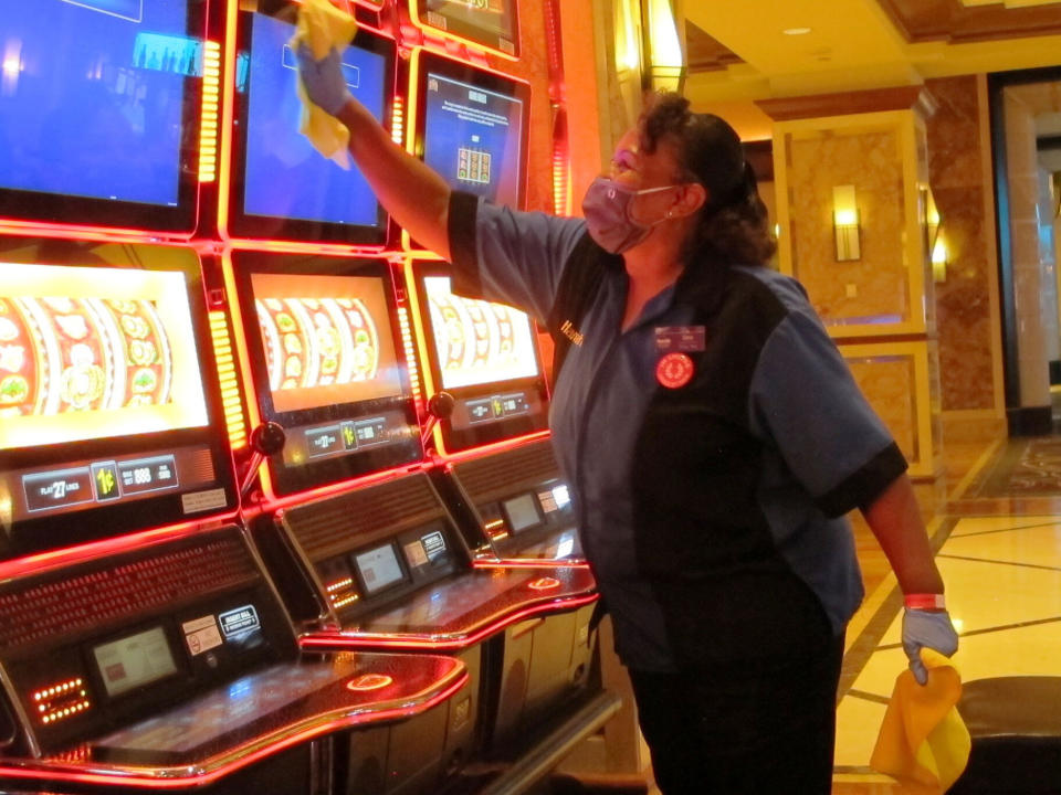 In this July 1, 2020 photo, Jeorganna Barnes, a worker at Harrah's casino in Atlantic City, N.J., wipes slot machines with disinfectant as the casino prepared to reopen after 3 1/2 months of being shut down due to the coronavirus. On Nov. 23, 2020, New Jersey gambling regulators released figures showing Atlantic City's casinos saw their gross operating profits decline by 37% in the third quarter of 2020 as they reopened under restrictions designed to slow the spread of the virus. (AP Photo/Wayne Parry)