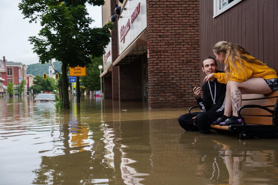 A man and a woman sit on a park bench with water up to the man's knees.  The woman is sitting on the chair back.  A car in the street is flooded up to the roof.