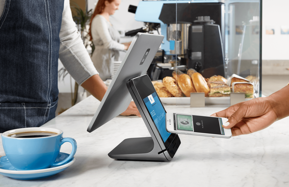 A hand holding a smartphone over a Square terminal at the counter of a cafe.