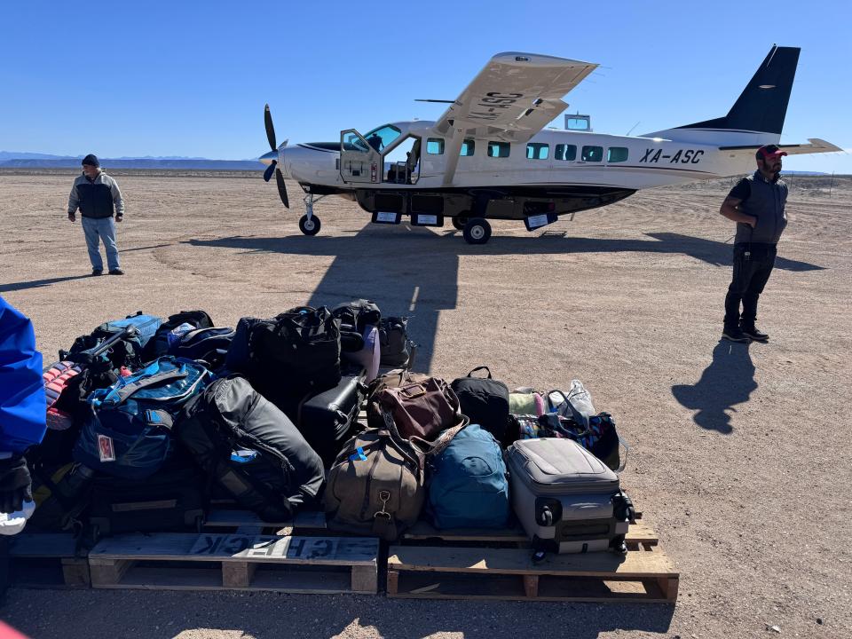 Luggage in a pile outside a small plane. Two men are standing near the plane.