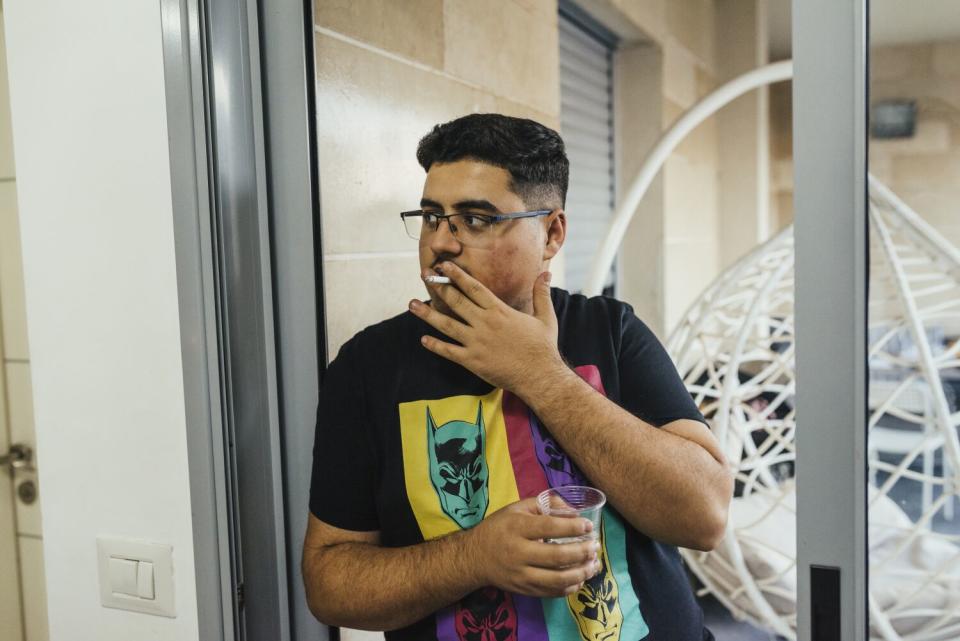 A man with dark hair, in glasses and a dark T-shirt, holds a cup in one hand and a cigarette in the other