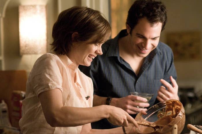 Amy Adams and Ron Livingston cooking together, laughing while she mixes batter in a bowl. Ron holds a drink, enjoying the moment in a kitchen setting