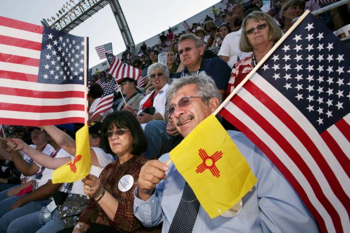 Un homme, assis dans des gradins avec une foule, tient les étoiles et les rayures et le drapeau rouge et jaune du Nouveau-Mexique