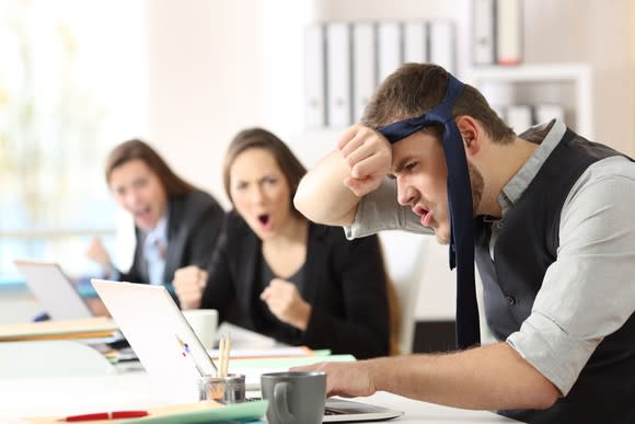 Young office worker with a necktie tied around his head, wiping his brow, working at his laptop, while two coworkers cheer his efforts in the background.