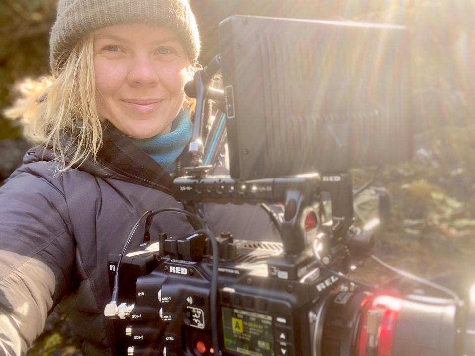 A selfie photo of Erin Ranny holding a camera with the sun shining above her. Erin has blonde hair and wears a brown hoodie and grey winter coat. She smiles slightly. 