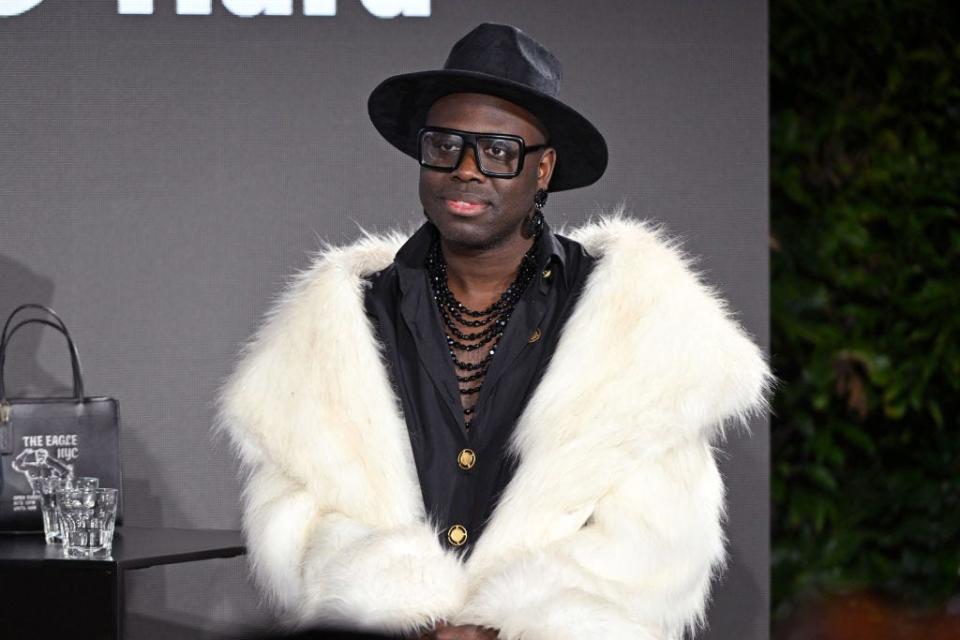 Bob the Drag queen wears a black wide-brimmed hat, glasses, a black button-down shirt and a furry white coat.