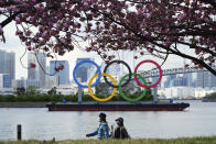 Under blooming cherry blossoms, people wearing protective masks to help curb the spread of the coronavirus walk with a backdrop of the Olympic rings floating in the water in the Odaiba section Thursday, April 8, 2021, in Tokyo. Many preparations are still up in the air as organizers try to figure out how to hold the postponed games in the middle of a pandemic. (AP Photo/Eugene Hoshiko)