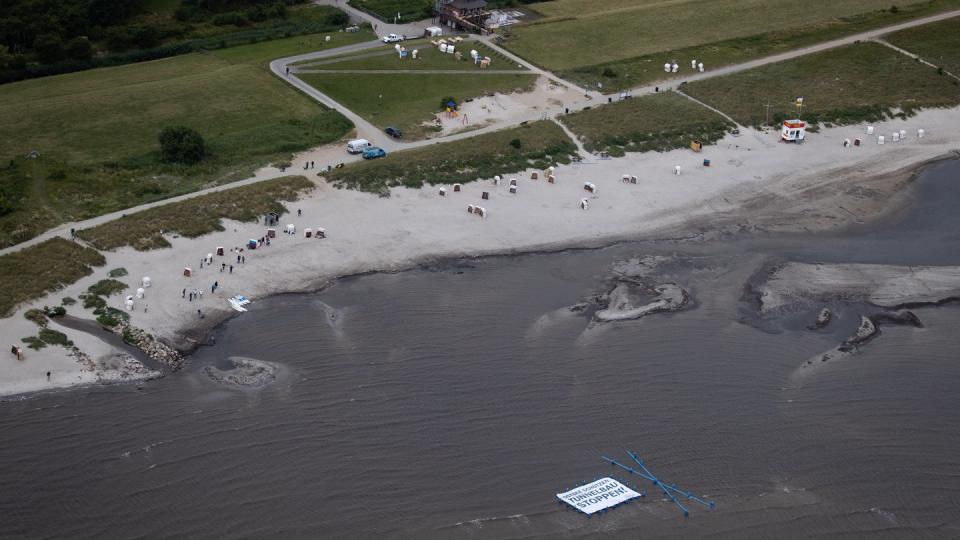 Unterstützer und Aktivisten der Protestaktion "Beltretter" halten ein Banner mit dem Text "Ostsee schützen. Tunnelbau stoppen!" und ein blaues Kreuz, während sie in der Ostsee am Naturschutzgebiet Grüner Brink nahe dem Fähranleger Puttgarden stehen.