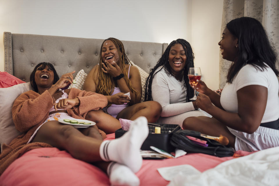 Four women laugh together during a relaxed gathering, seated casually with snacks and drinks