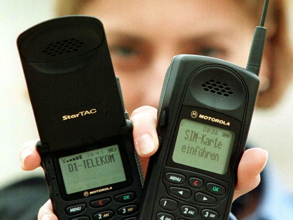 a blonde woman holds up a Motorola StarTAC flip phone and a slim motorola phone in one hand
