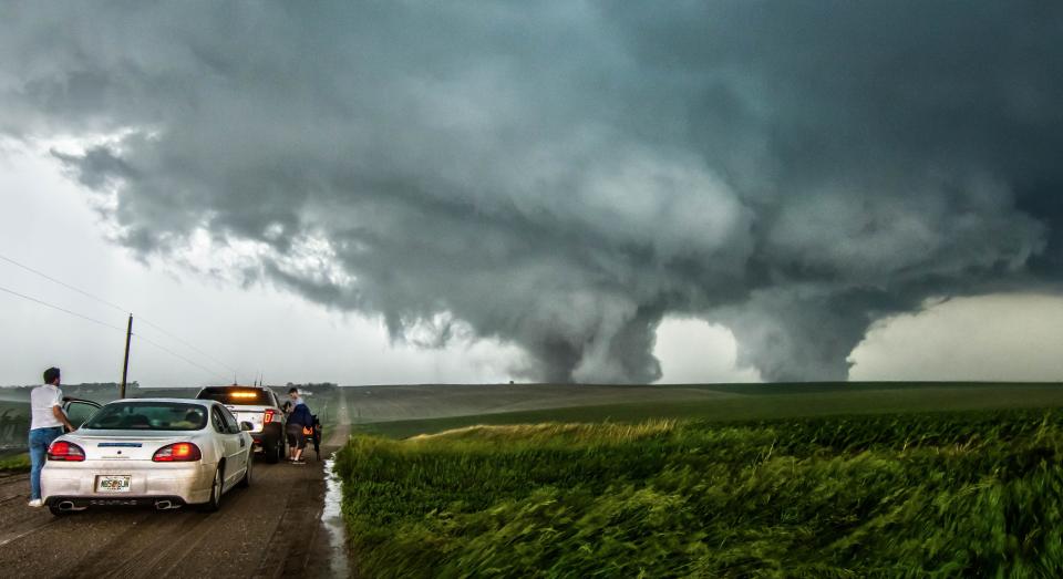Two tornados rage over a green field with two parked cars in the left foreground
