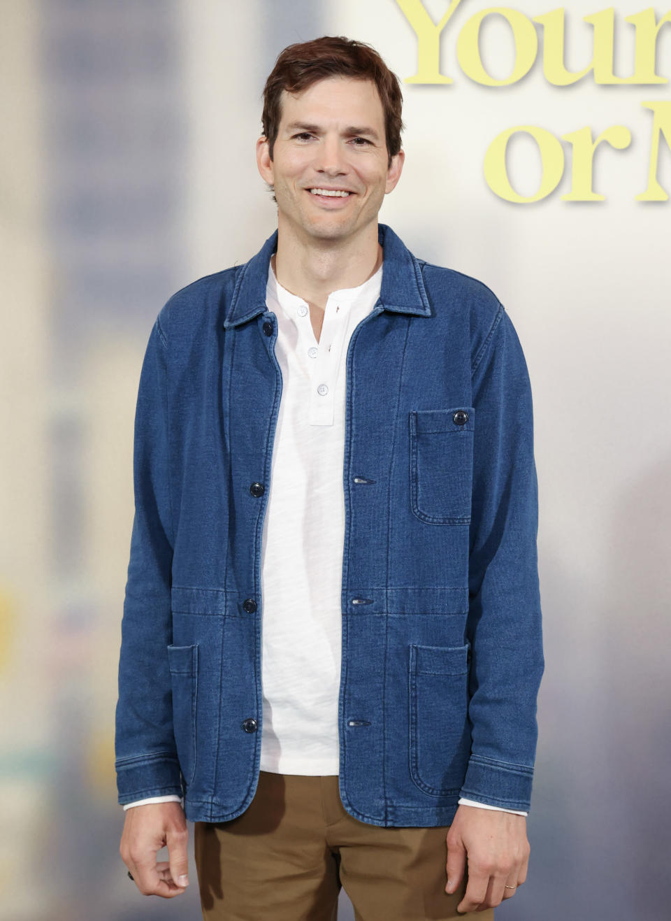 A close-up of Ashton smiling. He's wearing a t-shirt, a denim jacket, and slacks