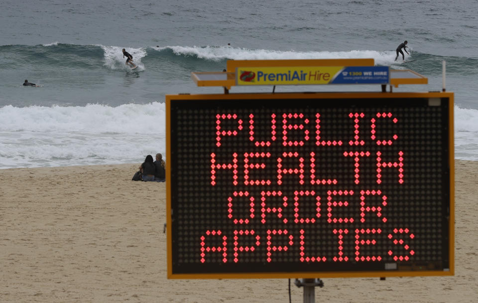 Surfers ride a wave past a sign at a beach in Sydney, Australia, Saturday, Dec. 19, 2020. Sydney's northern beaches will enter a lockdown similar to the one imposed during the start of the COVID-19 pandemic in March as a cluster of cases in the area increased to more than 40. (AP Photo/Mark Baker)