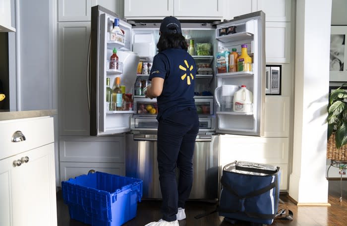 A woman wearing a Walmart employee uniform puts groceries inside a refrigerator at someone's home.