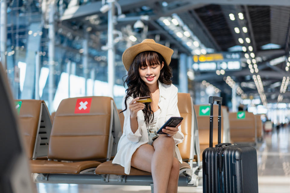 Asian young woman entering credit pin on the smartphone making hotel reservation while waiting for boarding in the airport terminal