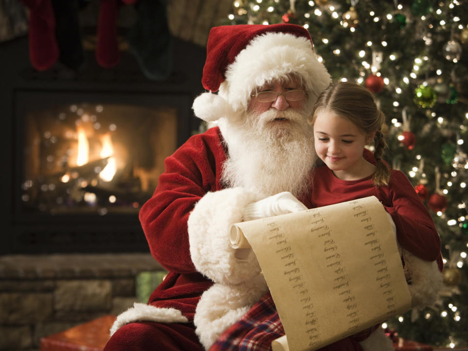 A child sits on Santa's lap in front of a Christmas tree and fireplace as Santa points to his list