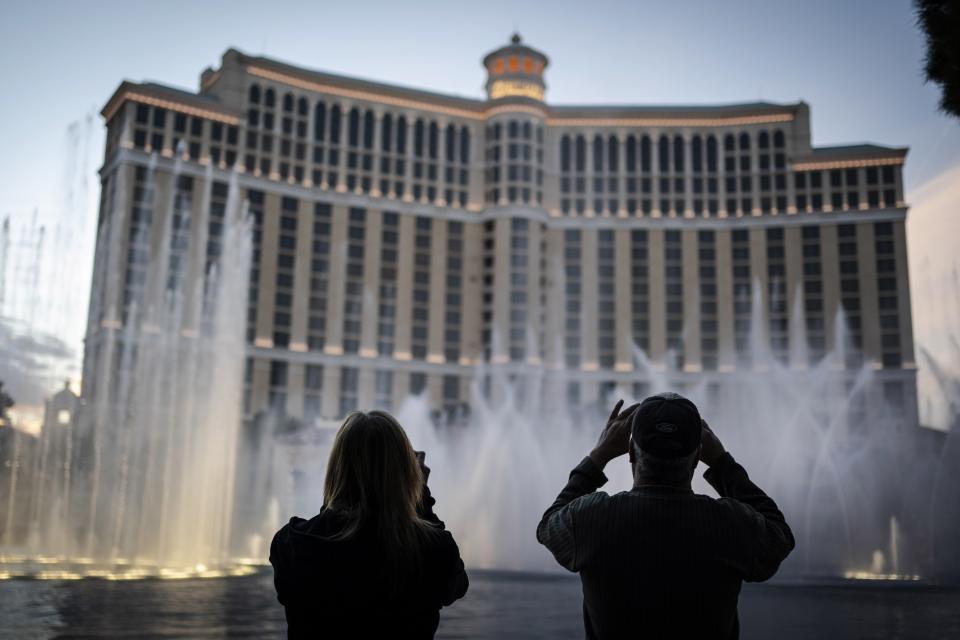 People are silhouetted as they take photos of the fountains at the Bellagio hotel-casino along the Las Vegas Strip, Wednesday, Nov. 11, 2020. Las Vegas sells itself on fantasies of wealth, luxury, and sex, and even the most cynical first-time visitor can come here expecting at least a hint of James Bond playing baccarat in Monte Carlo. That would be a mistake. Vegas feels more like a mixture of the endless mall and Disney-ish resort set to the music of amplified slot machines. Gamblers wear jeans and shorts, not tuxedoes. (AP Photo/Wong Maye-E)