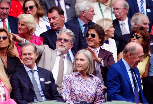 A man in a brown sits in the crowd with Tom Cruise, wearing dark sunglasses