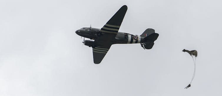 A paratrooper jumps from a second-world-war era plane against a grey sky.