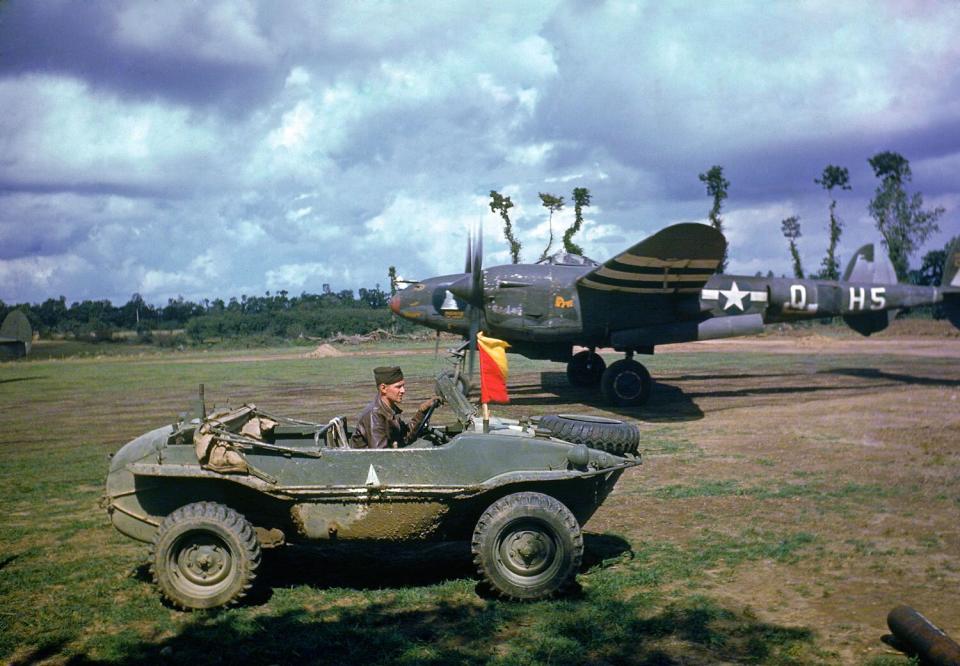 A P-38 fighter plane sits in the background as the pilot arrives in a captured German vehicle, France, 1944.