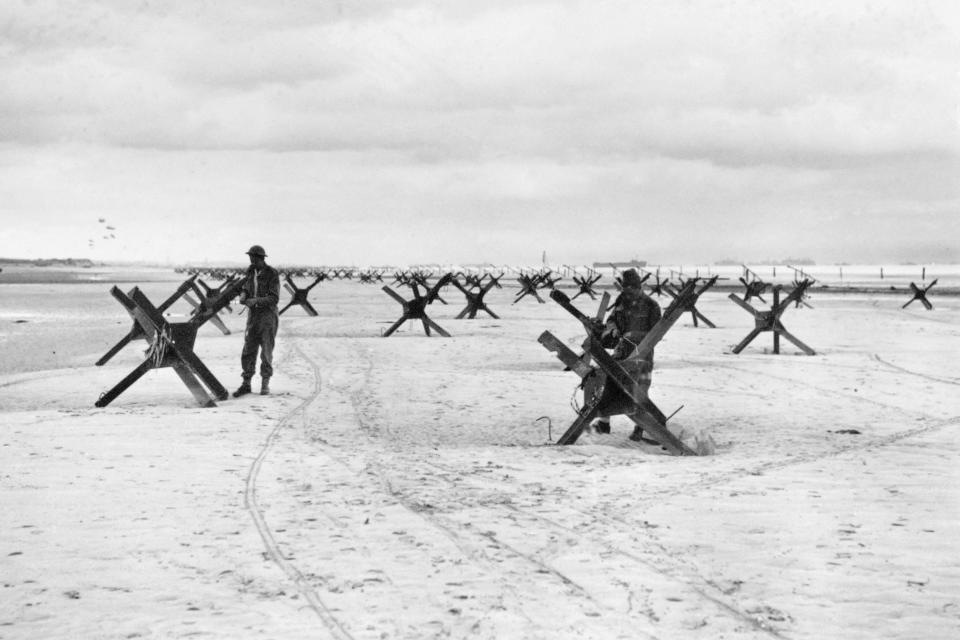 Black and white image of service members on a beach standing amid dozens of simple metal structures, each about 5 feet high.