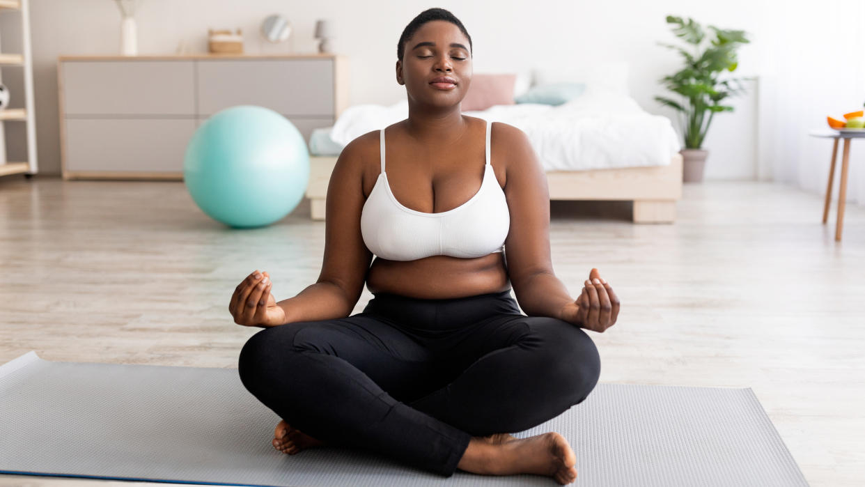  Woman in white tank top and black leggings sat meditating on yoga mat in living room. 