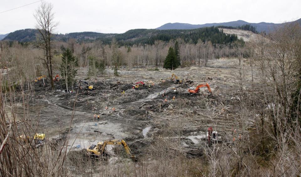 Workers clear debris and search at the scene of a deadly mudslide from the now-barren hillside at upper right, nearly two weeks earlier, Thursday, April 3, 2014, in Oso, Wash. More than a dozen people are listed as missing and 30 bodies have been found in debris from the March 22 landslide that broke off a steep hill, roared across the North Fork of the Stillaguamish River and buried a community at Oso, about 55 miles north of Seattle. (AP Photo/Elaine Thompson)