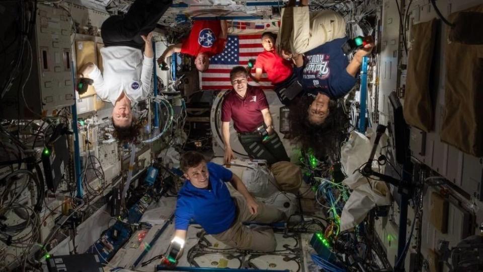 Mehrere Astronauten in einem dunklen Raumstationsmodul, deren Gesichter und Körper von Kamerabeleuchtung beleuchtet werden.  Auf der Rückseite ist eine amerikanische Flagge zu sehen