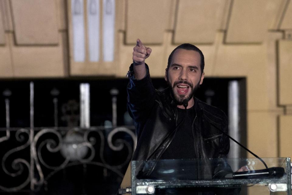 A man with a dark beard and wearing dark clothing speaks at a lectern while pointing with one hand