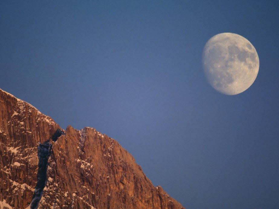 a picture shows the moon showing against the daylight sky behind a mountain ridge.
