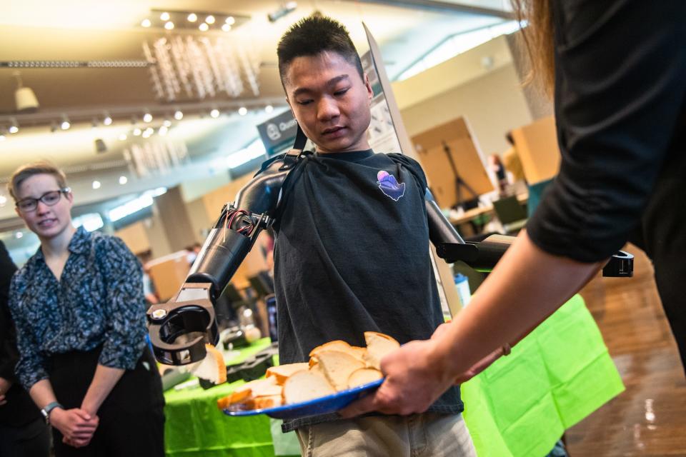 Colorado State student Jian Cohen uses prosthetic arms to pick up a piece of bread during an "Engineering Day," where senior undergraduate students showcase projects, on campus on Monday. A team of students designed the prosthetics that Cohen, who was born without arms, can use to eat and do other things more easily.
