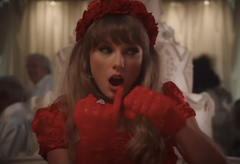 Taylor Swift in a red outfit and matching headpiece, holding a finger to her lips