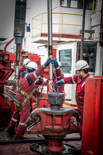 people in protective clothing and helmets on the deck of a ship, lifting a long metal pipe with water coming out of the bottom