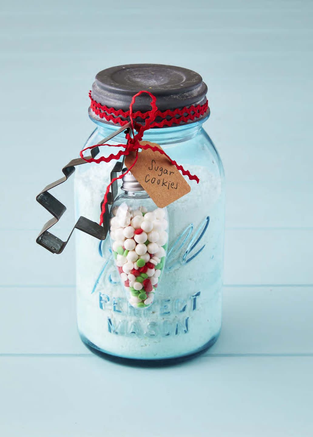 a vintage blue mason jar filled with dry sugar cookie mix the jar has a vintage tree shaped cookie cutter and light bulb shaped ornament attched to the outside