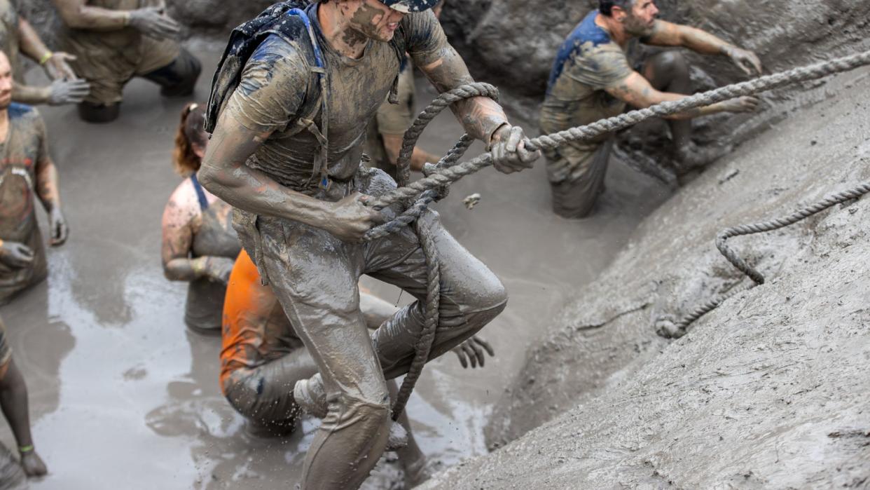  photo of a man coated in mud and holding a rope to climb up a wall in an obstacle course; others are pictured behind him, waiting in a pool of mud 