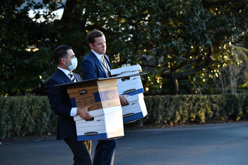 Two men wearing suits, and one wearing a face mask, each carry two cardboard boxes for files.