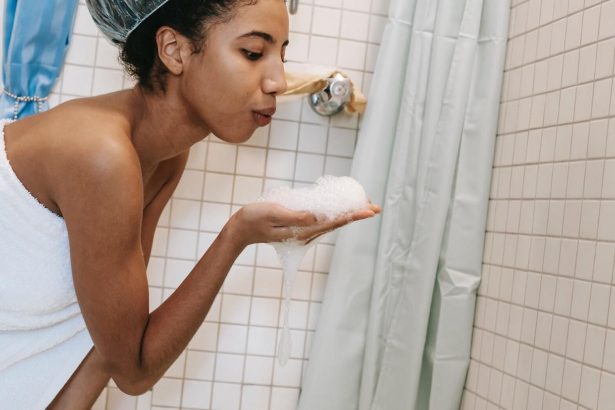 Black woman next to bath tub blowing bubbles out of hand in bathroom