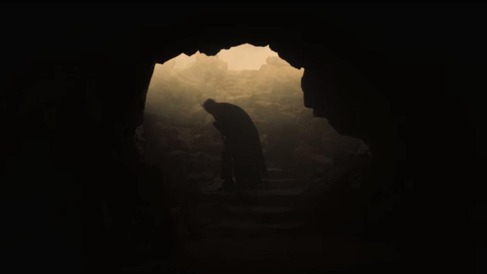 A man stands hunched over in the lighted mouth of a dark cave in the desert on Midnight Mass