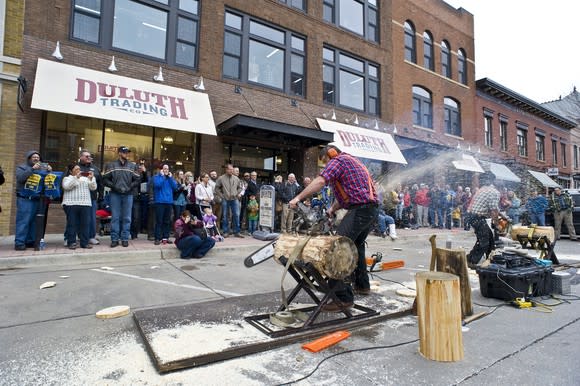 Person in a flannel shirt with a chainsaw cutting a log in front of a Duluth Trading store, with a modest audience of onlookers.