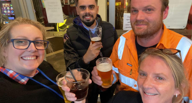 Four individuals, two women and two men, smile at the camera as they hold up their drinks at a pub amid public transport delays. 