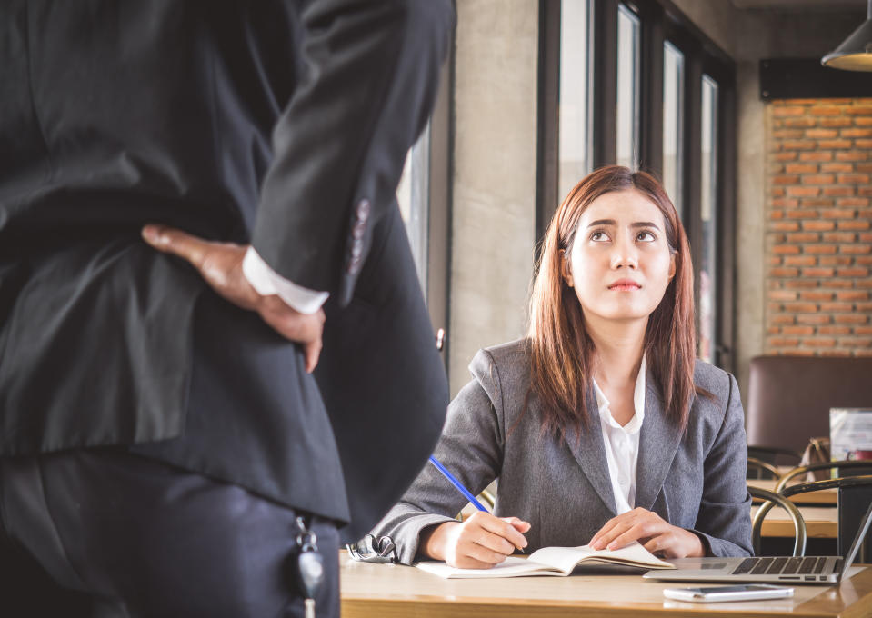 A person in the workplace sits at a desk with a notebook and pen in hand as they worriedly look up at their boss who has their hands on their hip