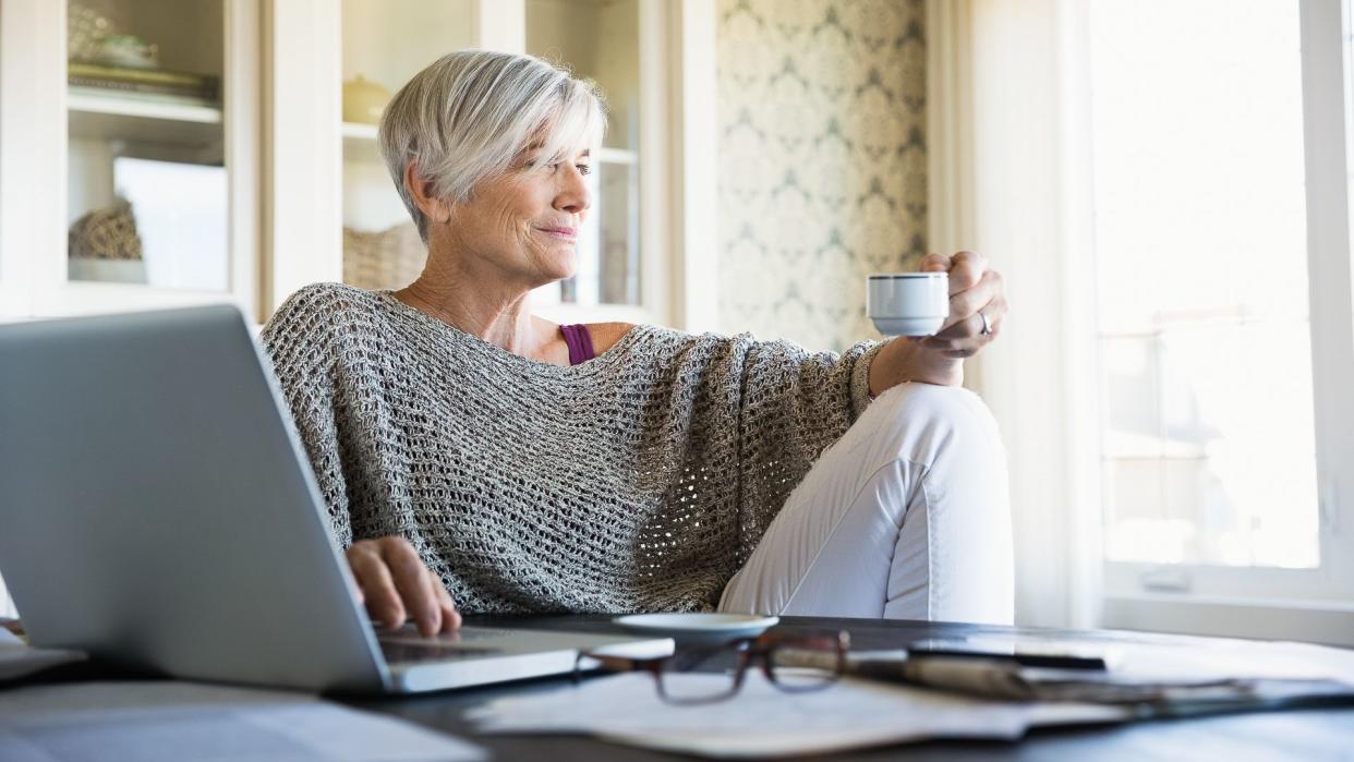 Pensive woman looking away drinking tea at laptop.