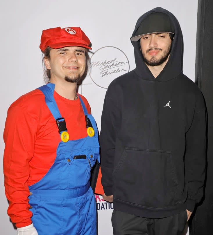 Person on left dressed as Mario, with overalls and cap, person on right in black hoodie and cap, both standing