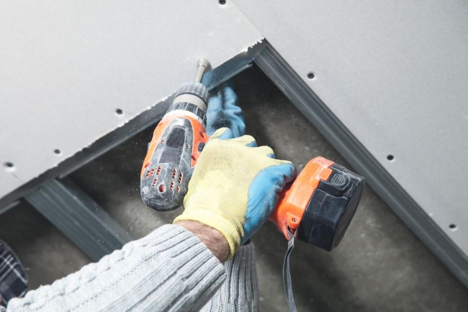 A close up of hands in yellow gloves using a drill to install a white ceiling. 