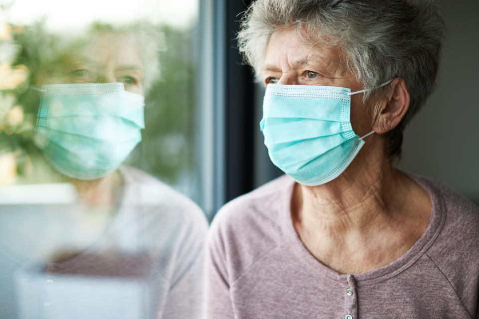 a old woman or grandma is wearing a respirator or surgical mask and is looking out the window while she is in quarantine because of the corona virus