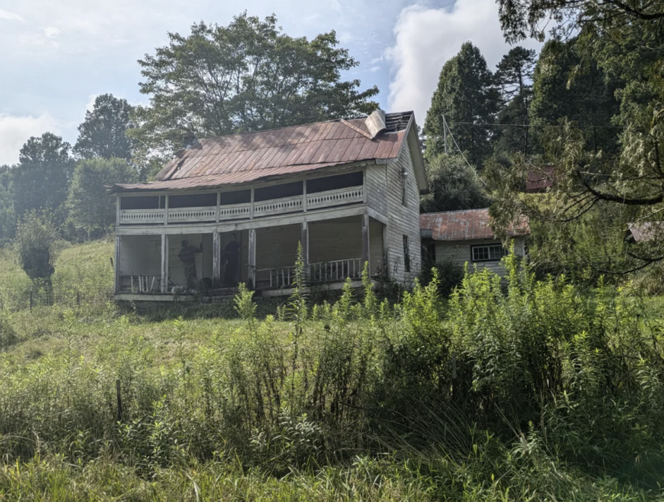 An old, weathered house stands in a lush, overgrown field with tall grass and surrounding trees in the background. The image evokes a feeling of abandonment