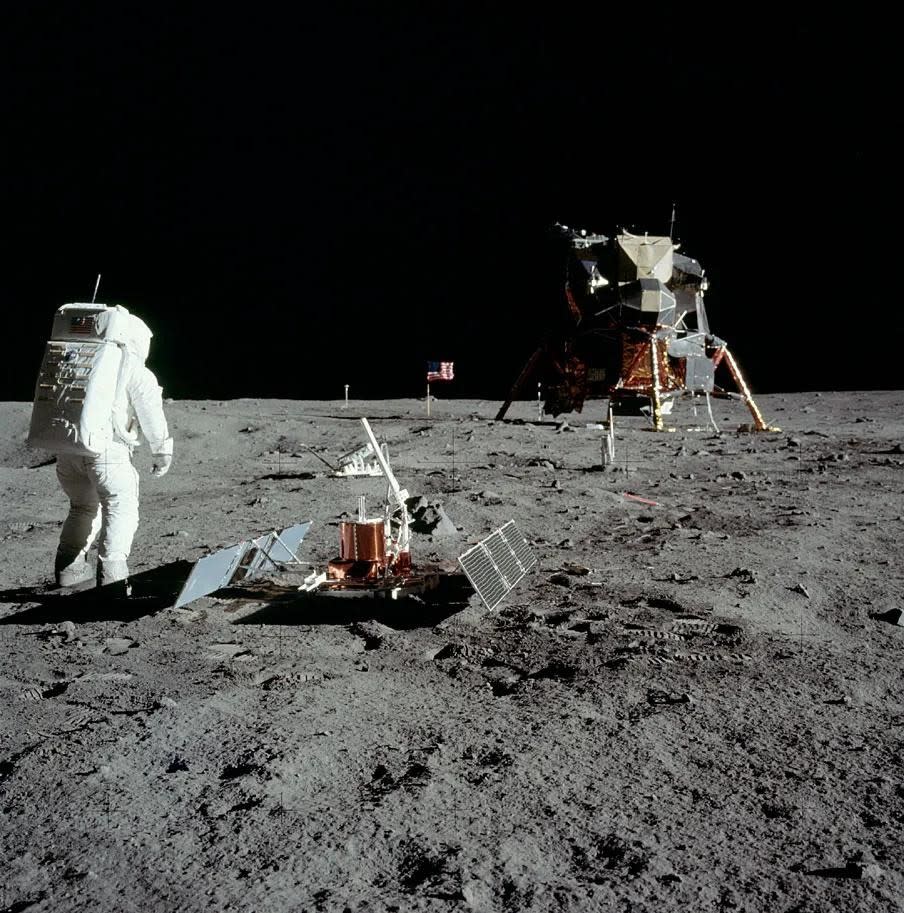 astronauts in bulky spacesuits walk across the gray dusty surface of the moon