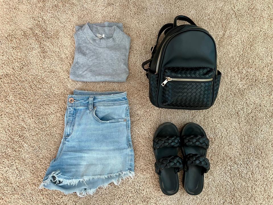 Grey shirt, denim shorts, black backpack, and black sandals spread out on floor.