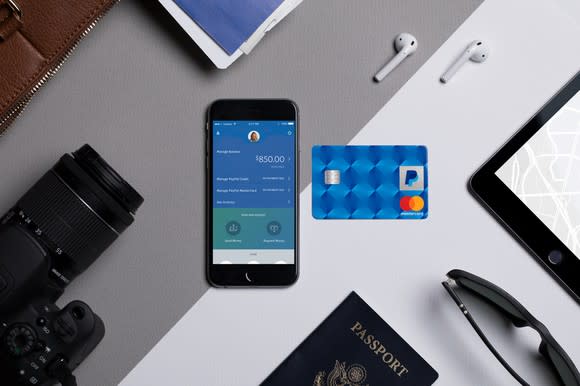 PayPal Cashback Mastercard surrounded by travel items: music ear buds, camera, mobile phone, passport, etc.